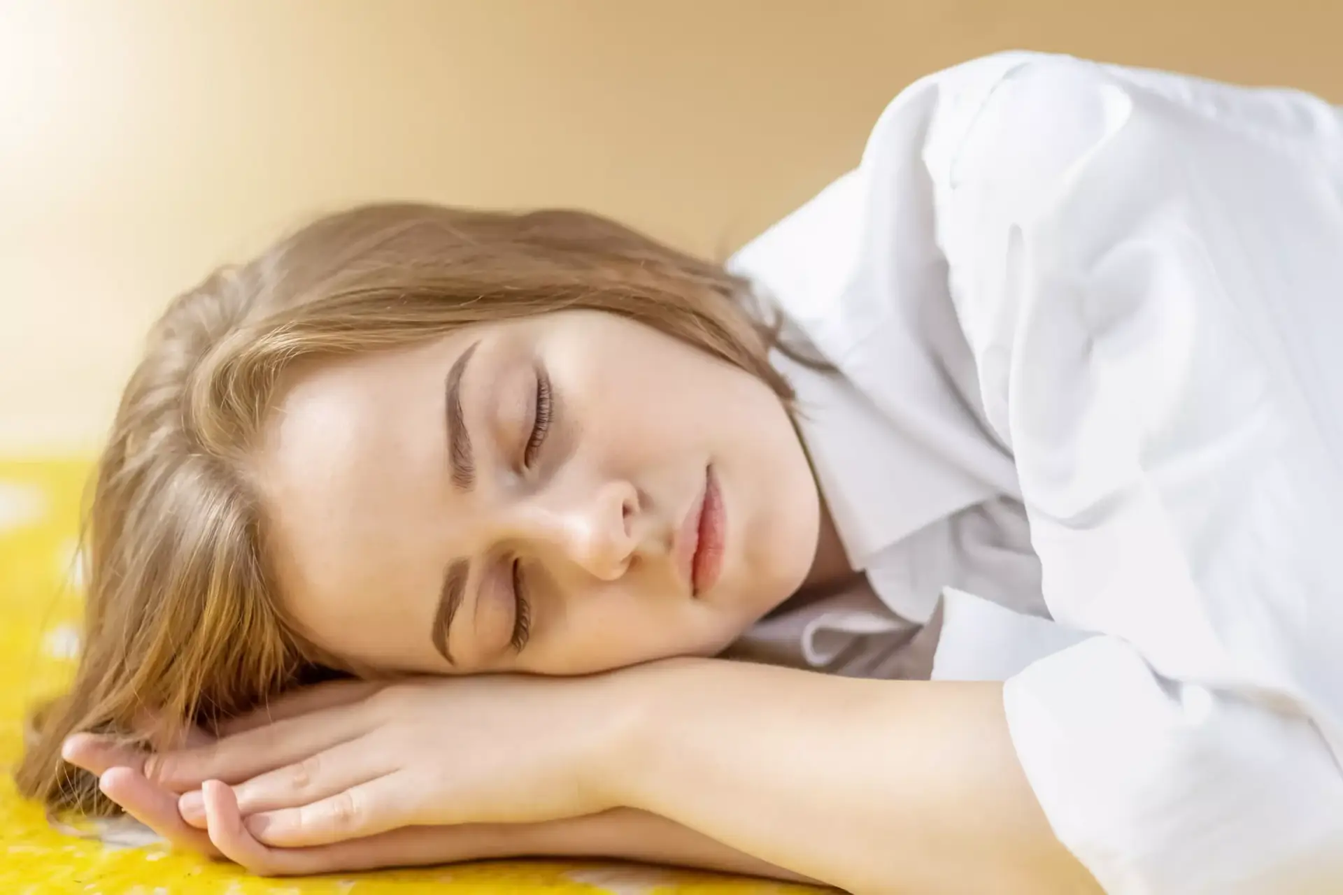 A young woman with light brown hair, wearing a white shirt, is lying on her side with her head resting on her hands, sleeping or resting on a yellow surface. STG-Health